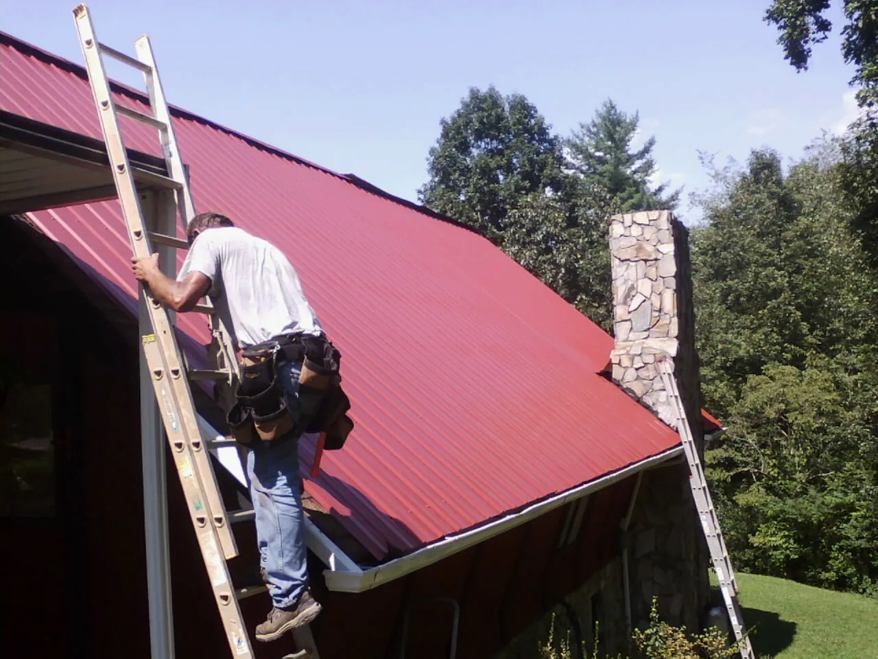 Roofer on a ladder beside a red metal roof and stone chimney — Soffit & Fascia Repair by Roofing Brothers in Bettsville